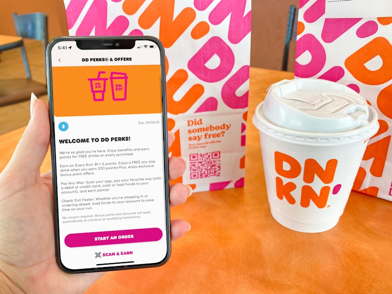 A person's hand holding an iPhone displaying the welcome to DDPerks page on the Dunkin app next to a small coffee and a Dunkin bag ...