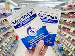 Two packages of Mucinex Extended Release Tablets held out by hand in front of a store aisle.