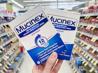 Two packages of Mucinex Extended Release Tablets held out by hand in front of a store aisle.