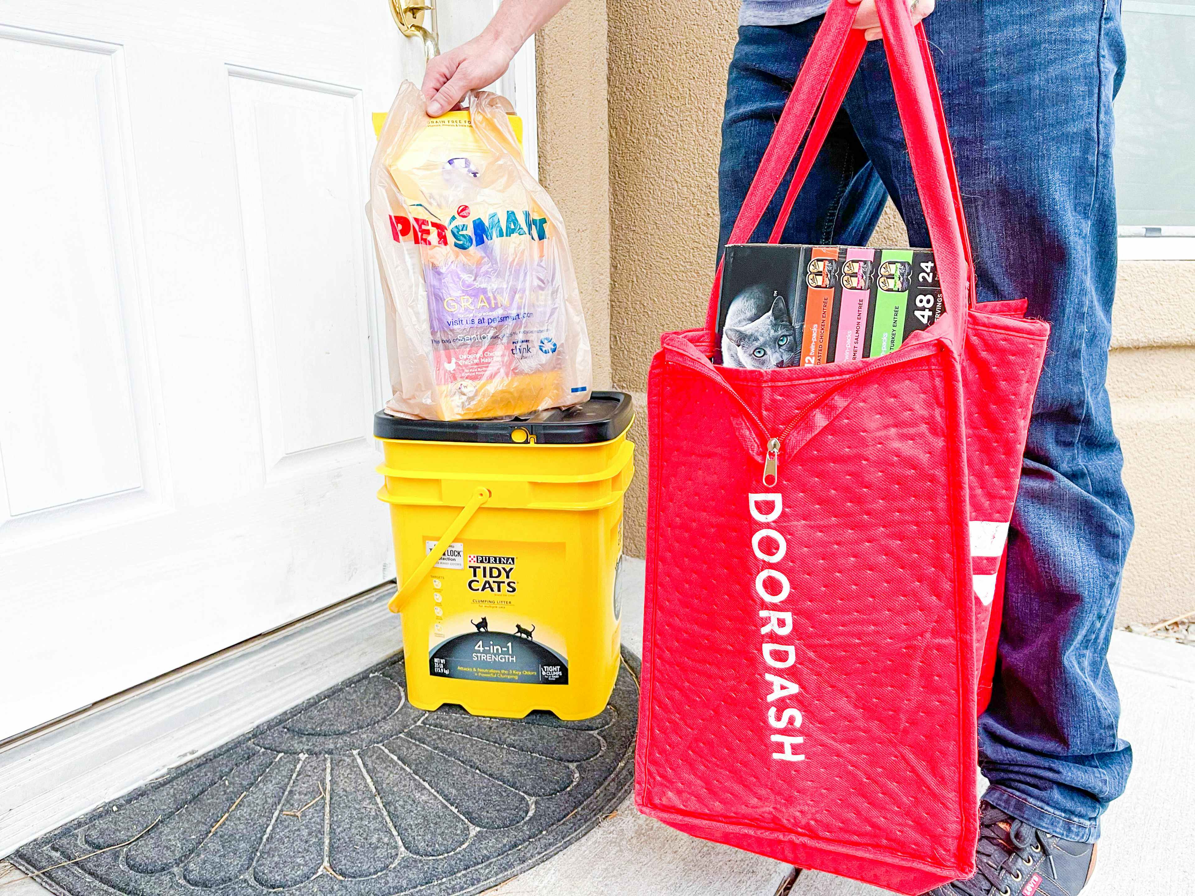 A Doordash delivery person standing on a front porch, setting a PetSmart bag onto a box of Tidy Cat kitty litter and holding a large Door...