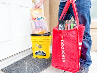 A Doordash delivery person standing on a front porch, setting a PetSmart bag onto a box of Tidy Cat kitty litter and holding a large Door...