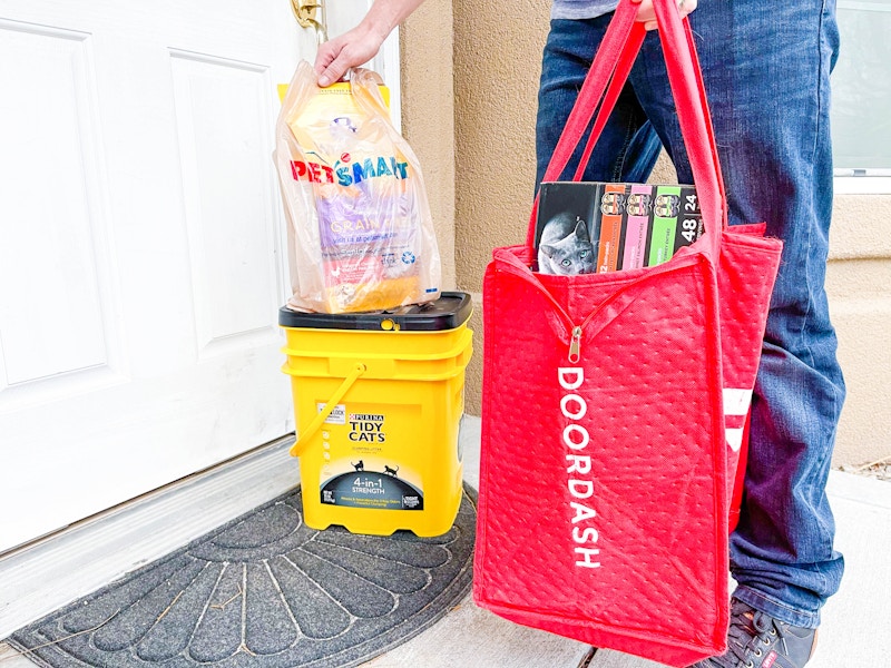 A Doordash delivery person standing on a front porch, setting a PetSmart bag onto a box of Tidy Cat kitty litter and holding a large Door...