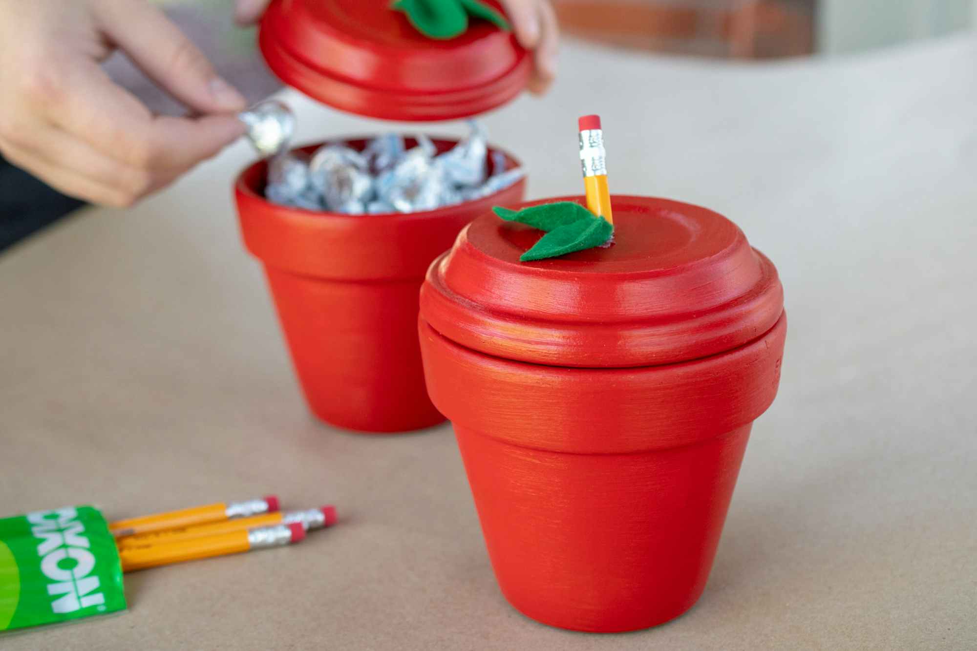 Person grabbing a small candy out of one if two small terra cotta pots painted red with a leaf and pencil end glued to the lids to ...