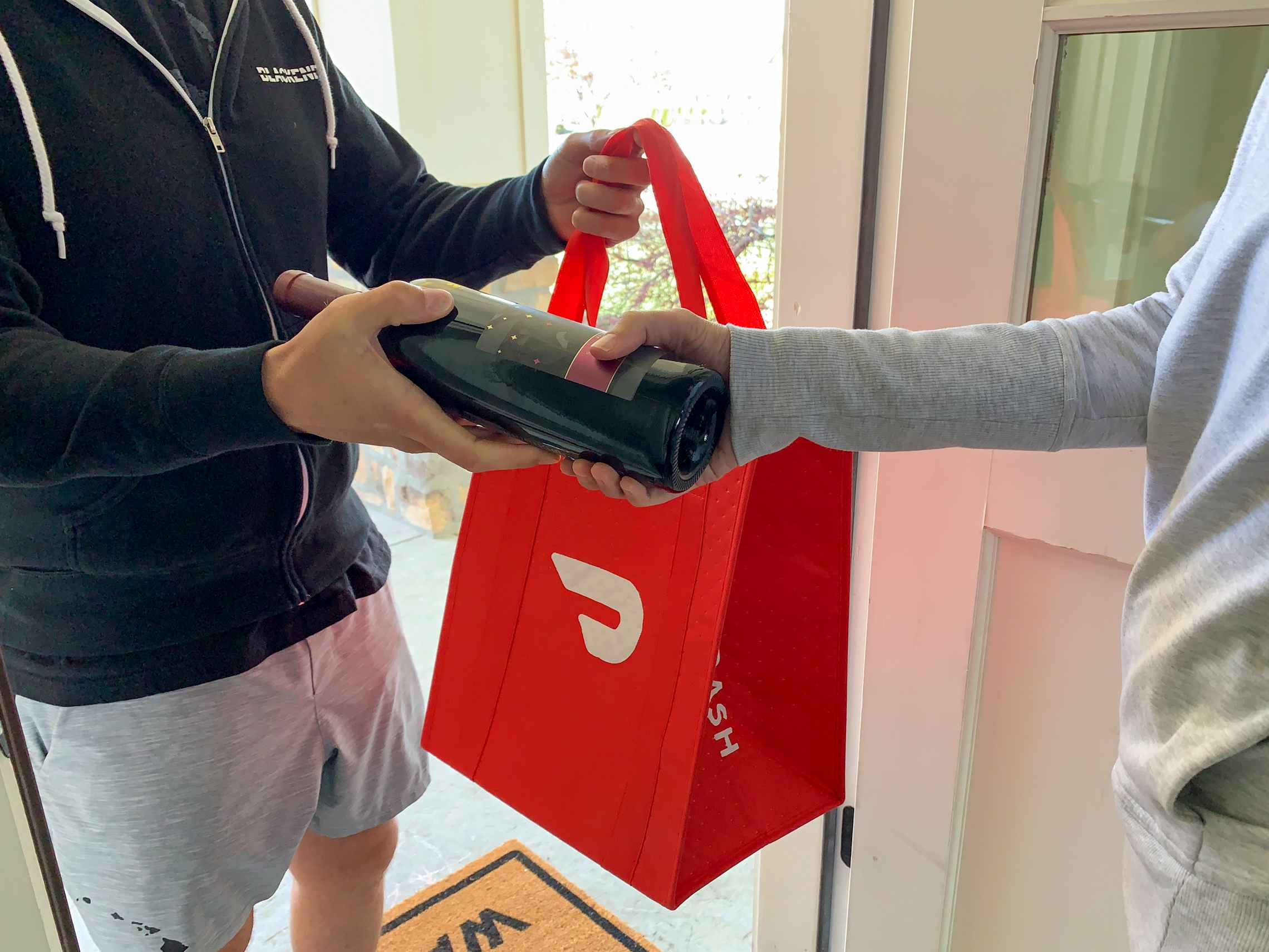 A Doordash employee holding a Doordash bag and handing a bottle of wine to someone in their front doorway.