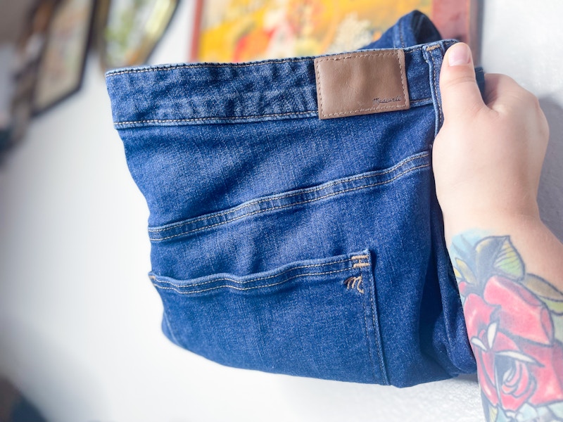 A person's hand holding up a pair of Madewell jeans in their home.