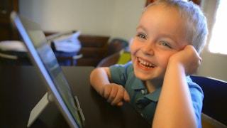 A small boy smiling at the camera with a tablet sitting in front of him