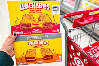 hand holding 2 boxes of lunchables grilled cheesies in front of a target cart