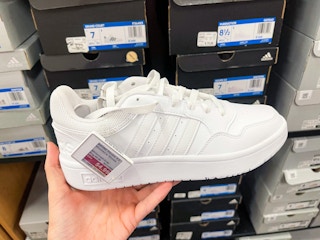 A white Adidas sneaker held out with hand in front of other Adidas boxes sitting on a store shelf.
