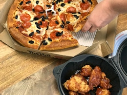 A person's hand taking a slice from a pizza sitting in a pizza box next to a container of wings.