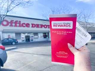 A person's hand holding up an Office Depot rewards brochure and a receipt outside of an Office Depot store.