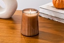 Lifestyle image of a Better Homes & Garden candle (scent: Farm Apple Pumpkin) on table with books/pumpkin 