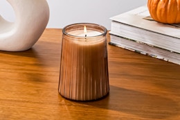 Lifestyle image of a Better Homes & Garden candle (scent: Farm Apple Pumpkin) on table with books/pumpkin 