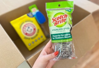 hand holding scotch-brite stainless steel scrubbers over a box with other houisehold items