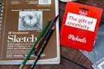 a michaels gift card next to a sketchbook and drawing pencils