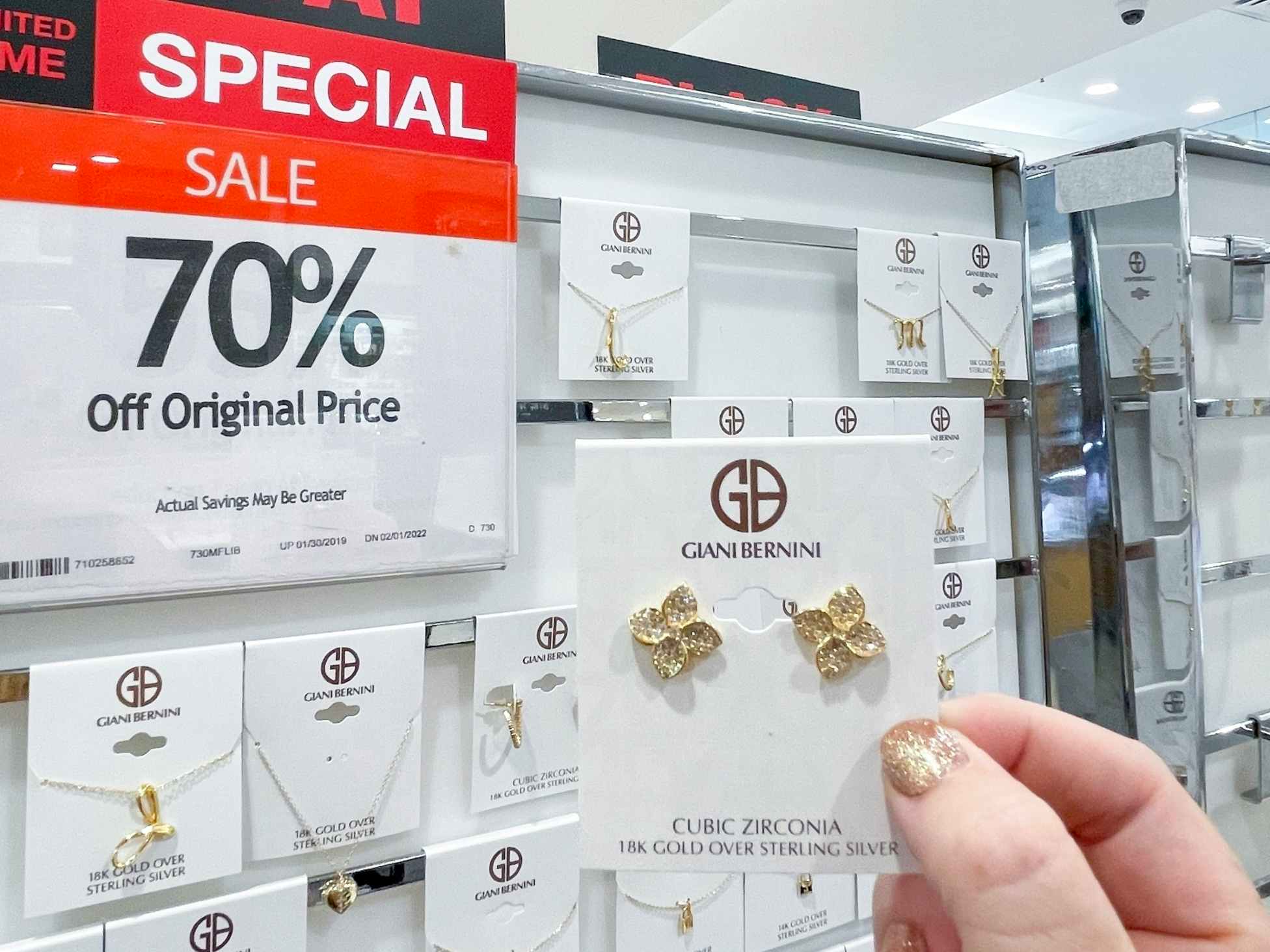 A person's hand holding up a pair of Giani Bernini earrings next to a jewelry display and a 70% off sale sign inside Macy's.
