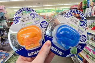 person holding 2 packs of gurunanda brush pods in an aisle