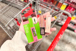 person holding maybelline. mascara, liquid eyeshadow, and lifter glaze balm over a cart