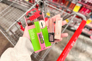 person holding maybelline. mascara, liquid eyeshadow, and lifter glaze balm over a cart