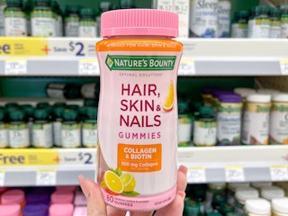 A bottle of Natures Bounty Hair, Skin, & Nails gummies held out by hand in front of a store shelf with other vitamin bottles on it.