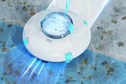 Round robotic pool cleaner with a clear window in the middle glides across several surfaces at the bottom of a pool.