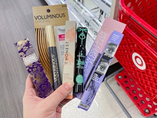 A person's hand holding a bunch of different brands of mascara next to a Target shopping cart.