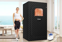 A woman stands in her living room in front of a big window onto the ocean with the black quilted sauna box beside her.