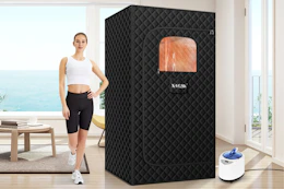 A woman stands in her living room in front of a big window onto the ocean with the black quilted sauna box beside her.