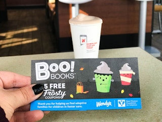 A person's hand holding a card that says, "Boo books 5 free jr frosty coupons" in front of a frosty on a table at Wendy's.