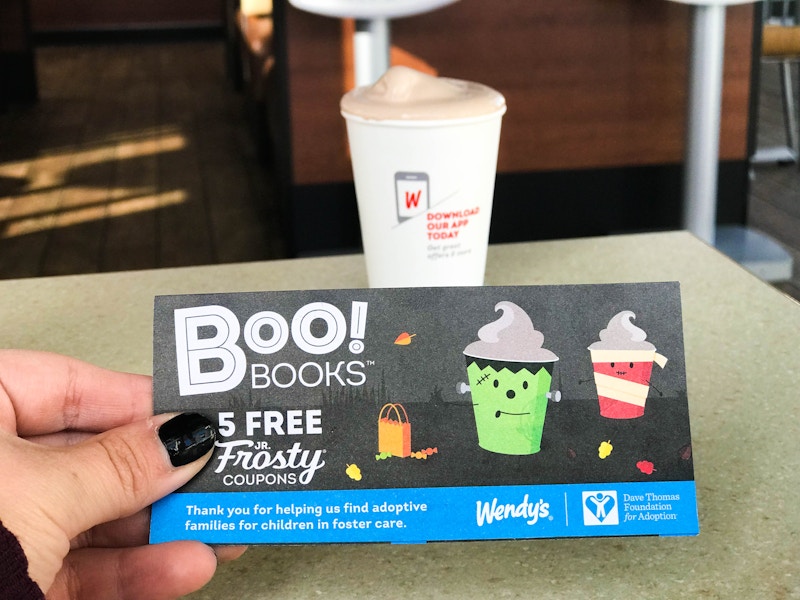 A person's hand holding a card that says, "Boo books 5 free jr frosty coupons" in front of a frosty on a table at Wendy's.