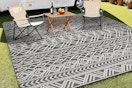A waterproof outdoor rug that's grey with a white line pattern has three lawn chairs on it. It sits on a grassy area by a trailer.
