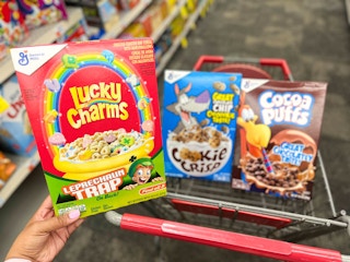 hand holding box of Lucky Charms with box of Cookie Crisp and Cocoa Puffs in cart