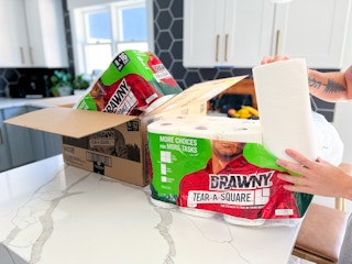 A hand holding up a paper towell roll in front of open Brawny packages