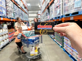 a person holding a costco card in front of a blurred image of a family shopping in costco
