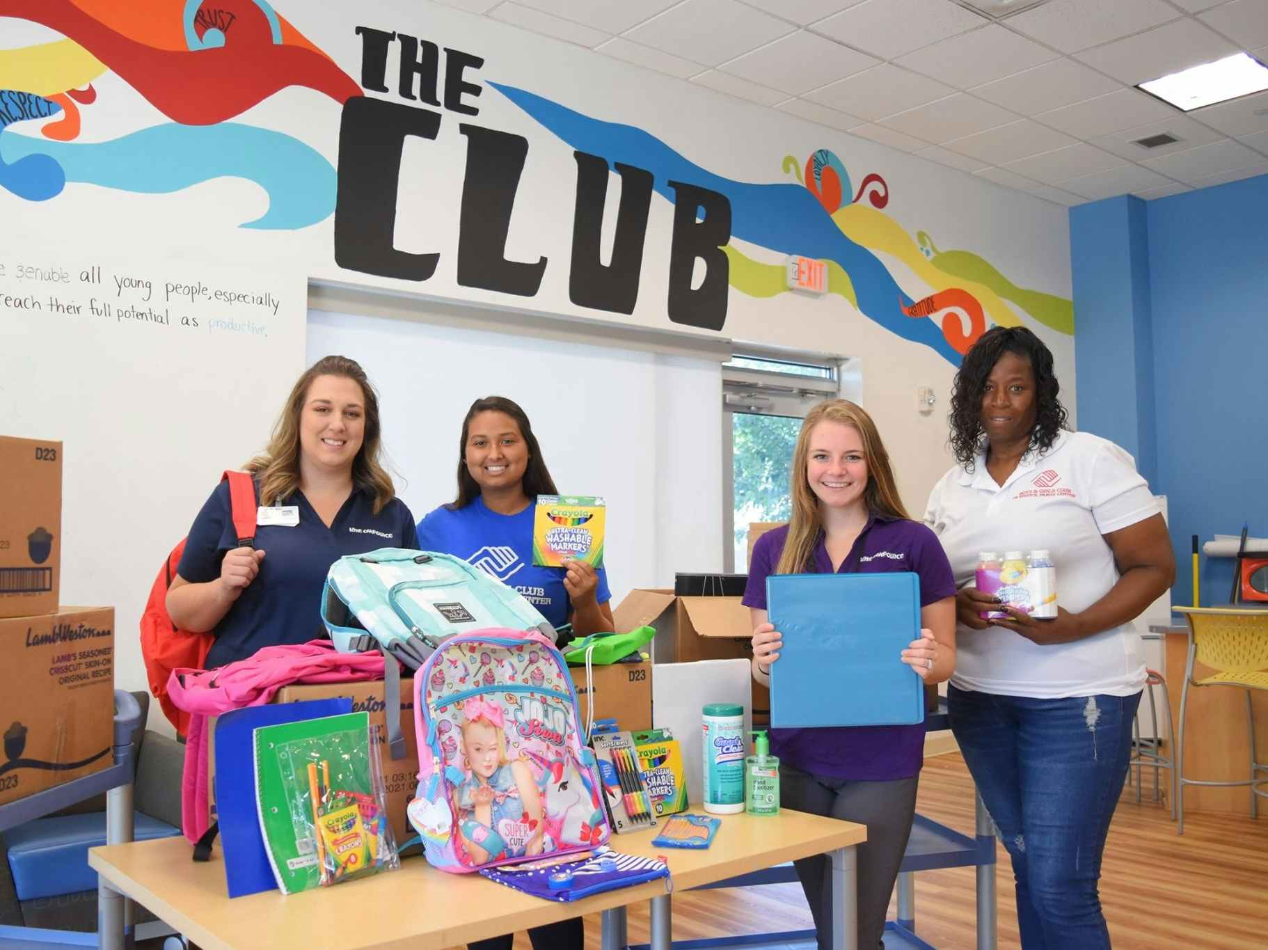 Three women posing for a photo with school supplies on a table in front of them.