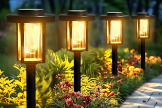 Four lit solar pathway lights illuminate a brick walkway and surrounding plants.