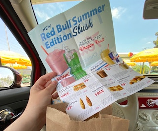 A person sitting in a car parked at Sonic, holding a page of Sonic coupons above a takeout bag.
