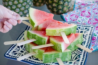 A person's hand taking a slice of watermelon on a popsicle stick from a plate of them.