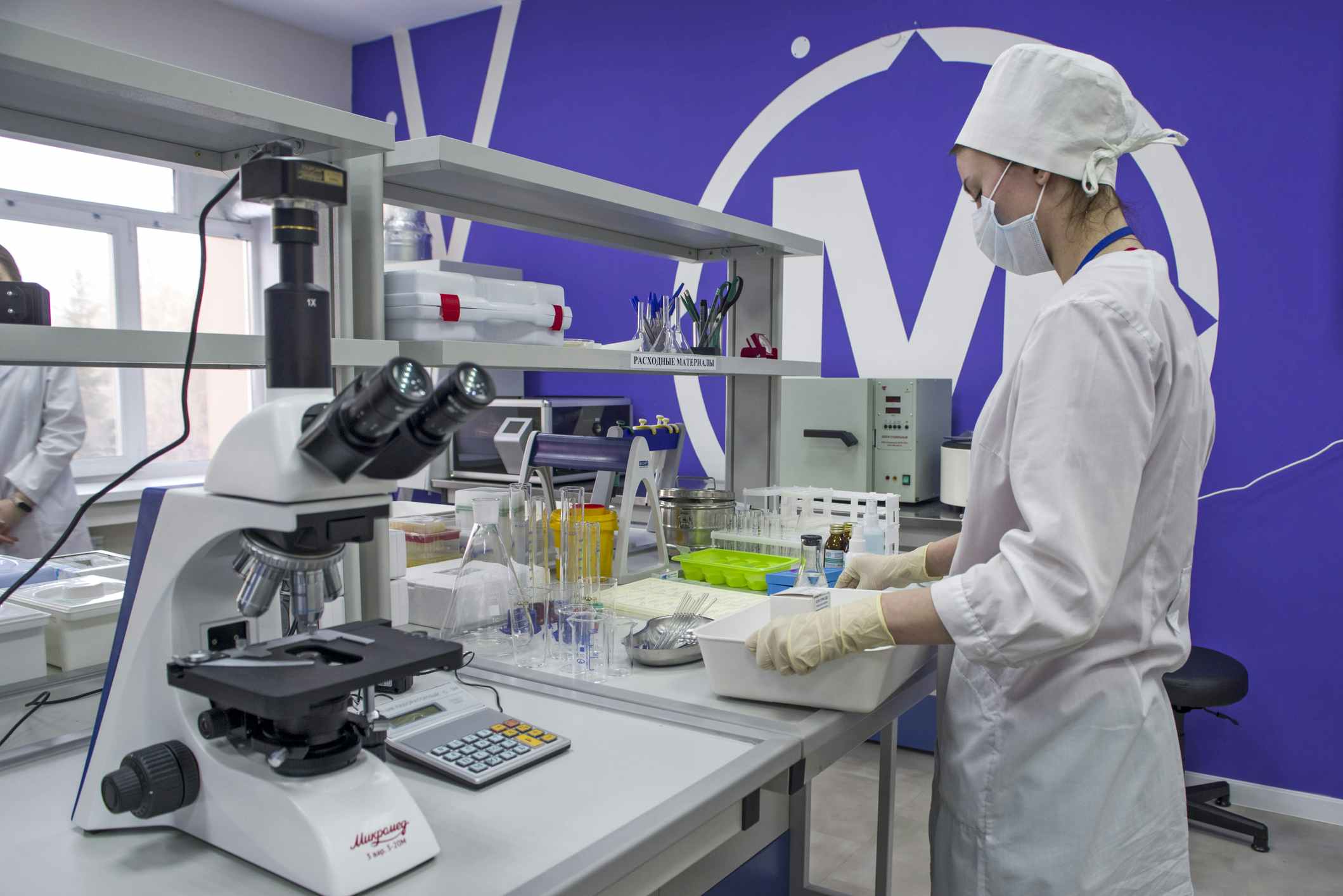 A medical student in a lab near a counter with microscopes and other medical lab equipment.