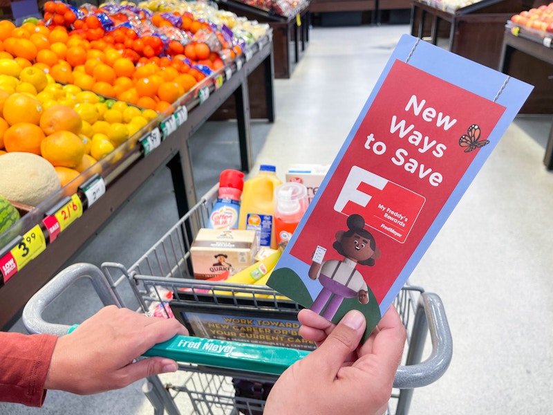 A person's hands, one holding a Fred Meyer rewards brochure, and the other pushing a Fred Meyer shopping cart full of groceries down the ...