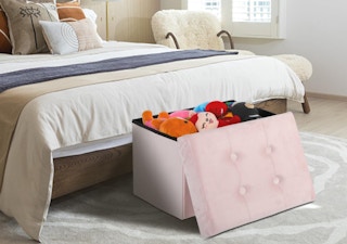 a pink ottoman filled with toys at the foot of the bed