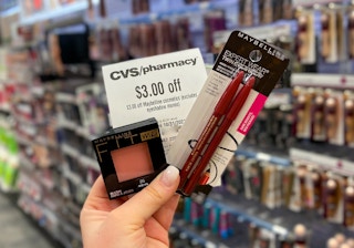 A person's hand holding up Maybelline makeup products with a coupon for $3 off at CVS.