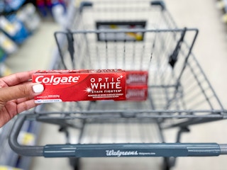 hand holding a box of Colgate Optic White stainfighter toothpaste in front of shopping cart with two boxes inside