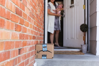 A mother holding her baby, opening the front door and looking down at an Amazon package sitting on her front porch.