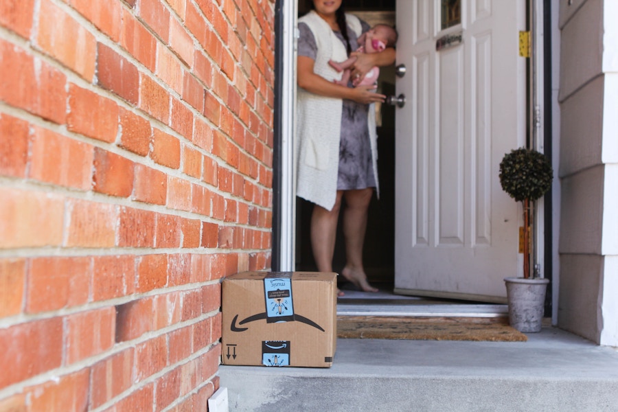 A mother holding her baby, opening the front door and looking down at an Amazon package sitting on her front porch.