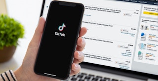 a hand holding an iPhone displaying the start up screen for the TikTok app in front of a laptop displaying an Amazon shopping cart ...
