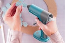 One hand holds a teal glue gun, the other holds a flower shaped polka dot piece of paper about to be glued to a cardboard circle.