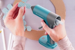 One hand holds a teal glue gun, the other holds a flower shaped polka dot piece of paper about to be glued to a cardboard circle.