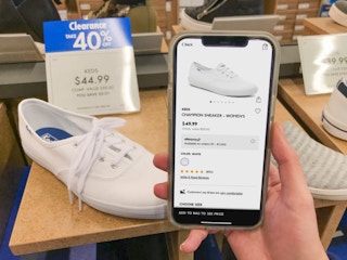 A white shoe on display with blue sales tag reading "40% off". A cell phone is held with the same shoe on the DSW app, with a regular price.