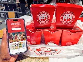 Someone holding a phone displaying the Panda Express app's main page featuring the family style meal next to some takeout boxes of food ...