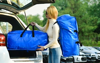 a woman carrying two big blue moving bags to her car's truck